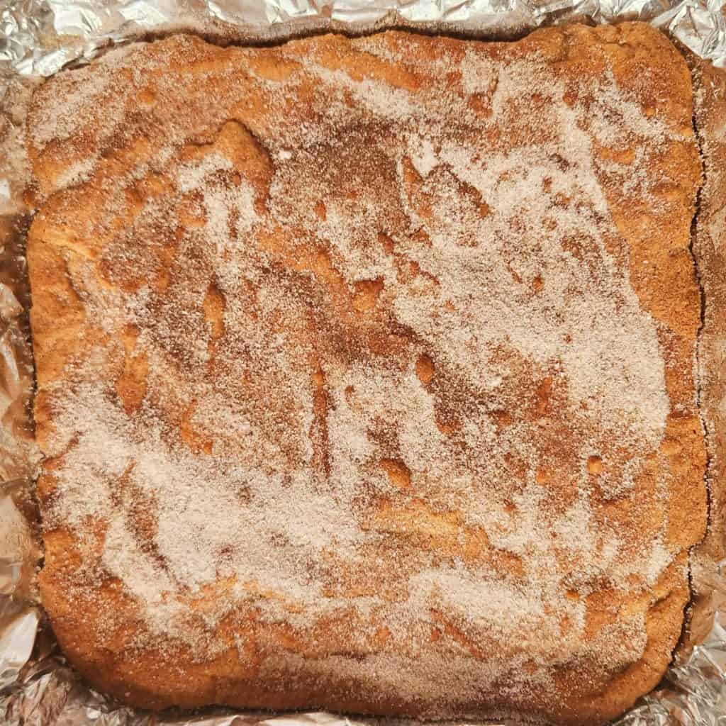 A baked square dessert covered with a layer of cinnamon sugar, set in a foil-lined pan.