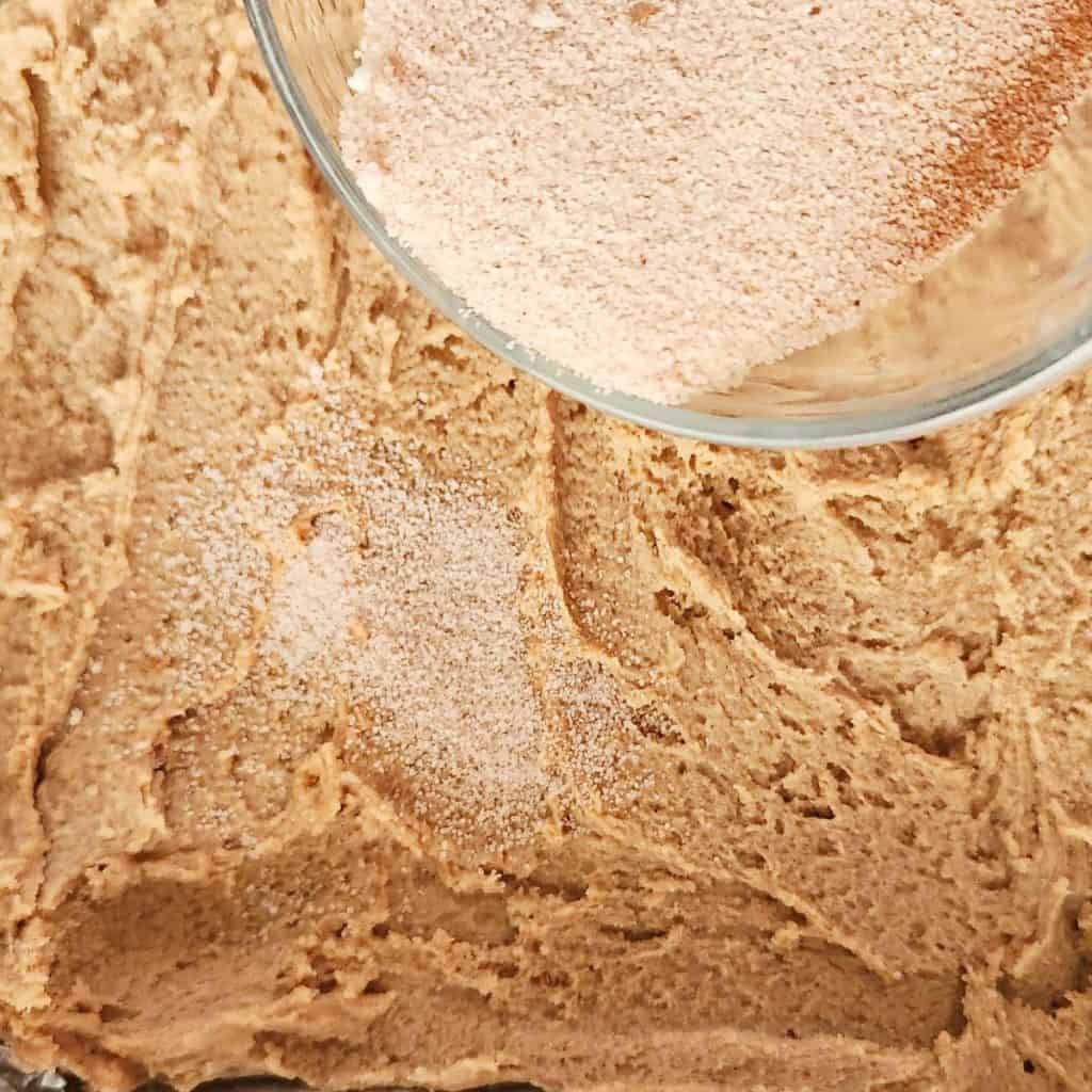 A glass bowl of cinnamon sugar is being sprinkled over a layer of cinnamon cookie dough.