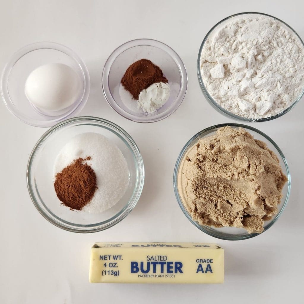 Top-down view of baking ingredients: one egg, flour, baking soda with cinnamon, granulated sugar with cinnamon, brown sugar, and a stick of salted butter on a white surface, ingredients for my baked churros bars recipe.