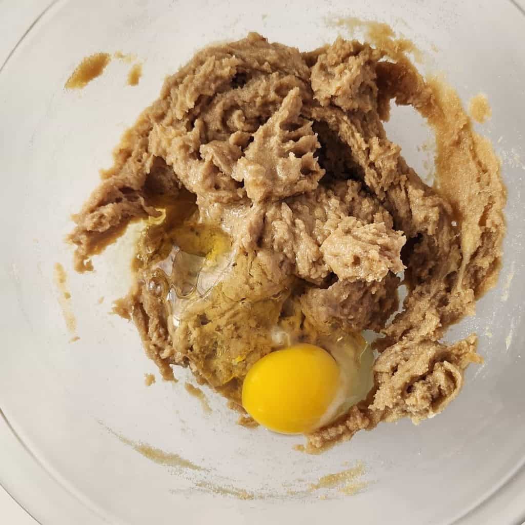 A glass bowl containing creamed brown sugar and butter with a raw egg cracked on top, ready to be mixed.