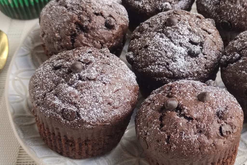 A plate of six chocolate zucchini muffins topped with powdered sugar and chocolate chips, displayed on a patterned plate.