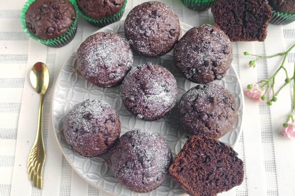 A plate of chocolate muffins topped with powdered sugar is surrounded by more muffins on a white tablecloth. One muffin is cut in half to show the inside. A gold spoon and a small pink flower are placed beside the plate.