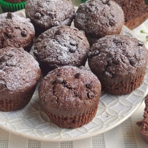 A plate of chocolate muffins topped with powdered sugar and chocolate chips sits on a white patterned tablecloth. Some muffins are whole, while a few are cut in half, revealing the moist chocolate interior.