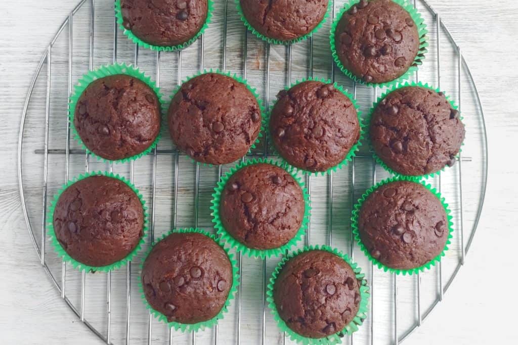 Chocolate muffins in green paper liners are arranged on a round cooling rack placed on a light-colored wooden surface.