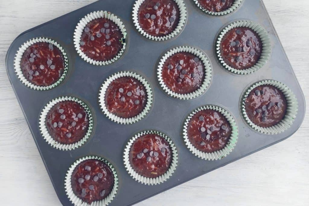 A muffin tin filled with 12 paper liners, each holding chocolate muffin batter topped with chocolate chips, is placed on a light-colored surface.