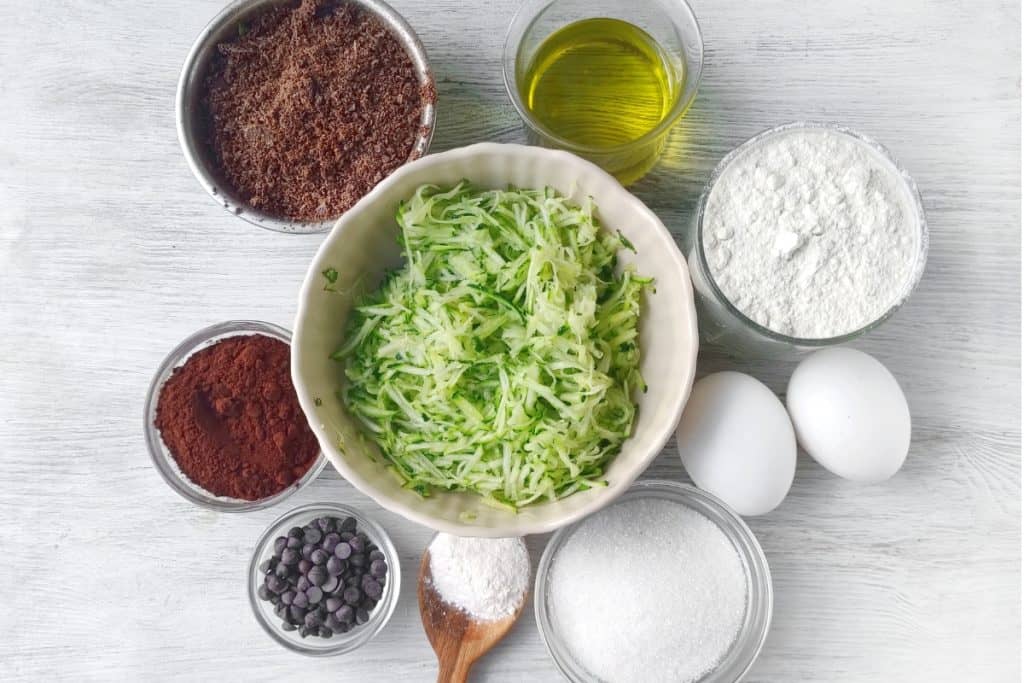 Ingredients for double chocolate zucchini muffins recipe, including a bowl of shredded zucchini is surrounded by ingredients in small bowls, including flour, cocoa powder, sugar, ground flaxseed, chocolate chips, a glass of oil, a spoonful of baking powder, and two eggs on a white wooden surface.