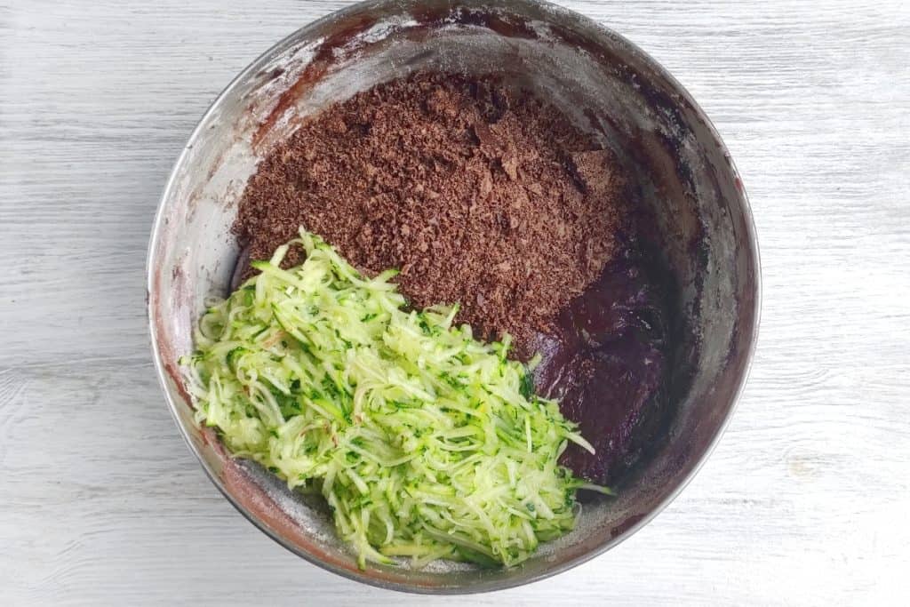 A metal mixing bowl containing shredded zucchini, cocoa powder, and chocolate cake batter on a light wooden surface.