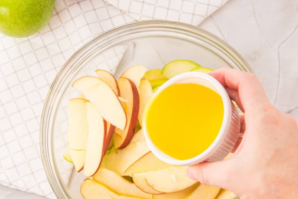 A hand holds a small white bowl of melted butter over a glass bowl filled with sliced apples, with a green apple and a checkered towel in the background.