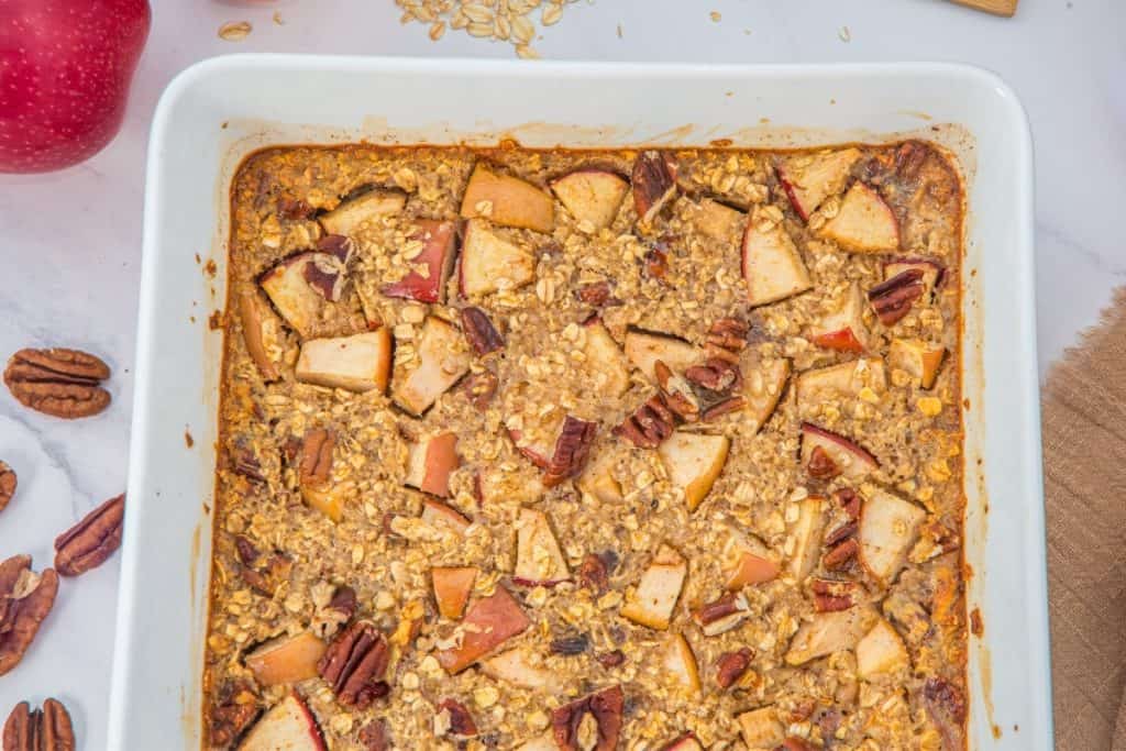 A baked oatmeal casserole with chopped apples, oats, and pecans in a white dish. Whole pecans and an apple are visible on the surface nearby.