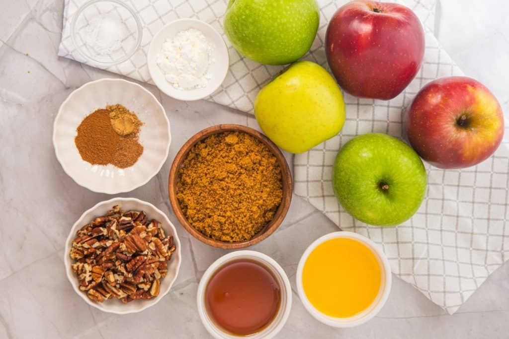 Four apples, bowls of brown sugar, chopped pecans, spices, cornstarch, and salt, plus honey and melted butter, are arranged on a white cloth and marble surface, showing ingredients for a baking recipe.