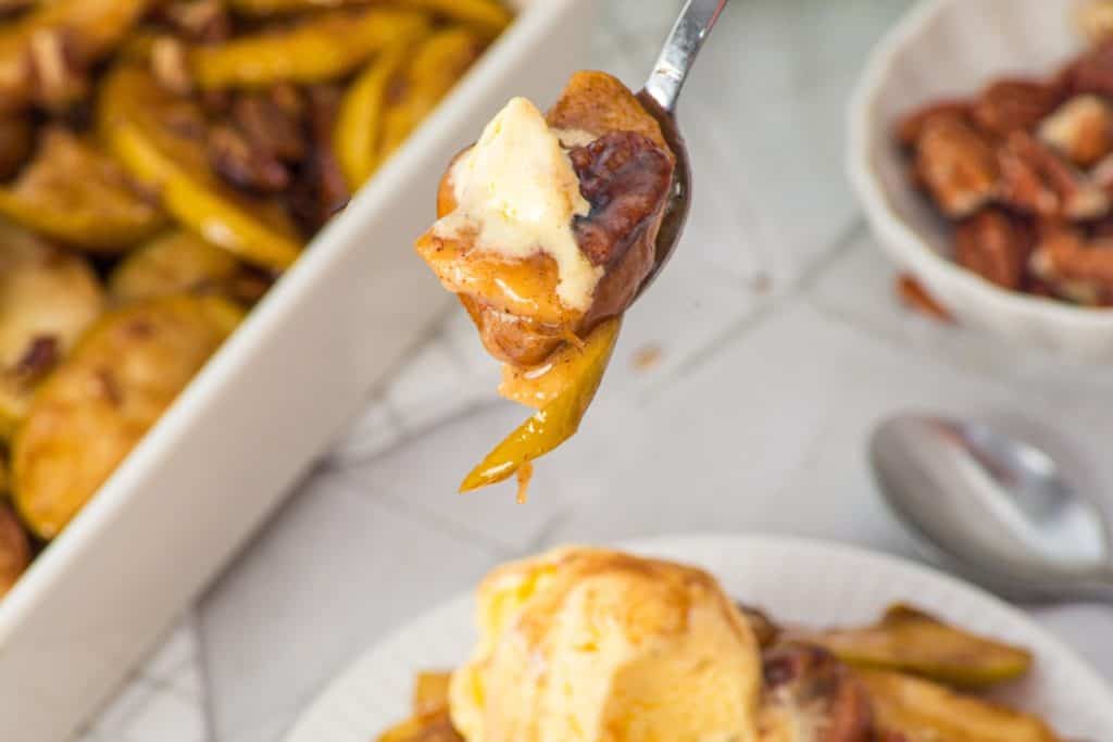 A spoon holds a bite of baked apples, pecans, and melting vanilla ice cream above a plate and dish of the same dessert.