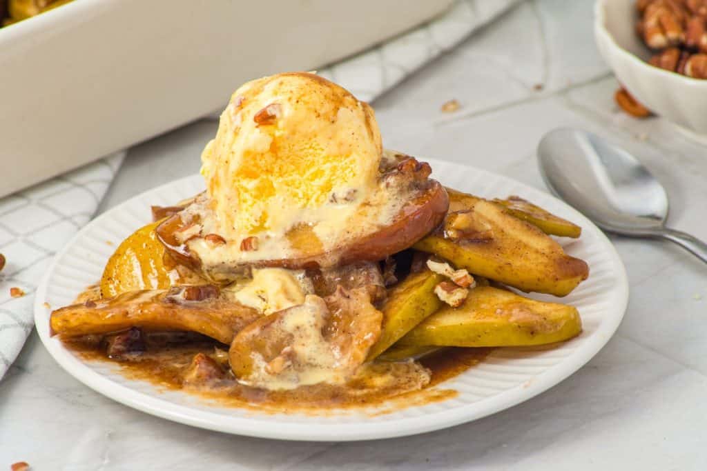 A plate of baked apple slices topped with a scoop of melting vanilla ice cream and sprinkled with chopped pecans. A spoon rests next to the plate on a white surface.