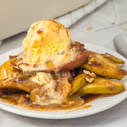 A plate of baked apple slices topped with a scoop of melting vanilla ice cream and sprinkled with chopped pecans. A spoon rests next to the plate on a white surface.