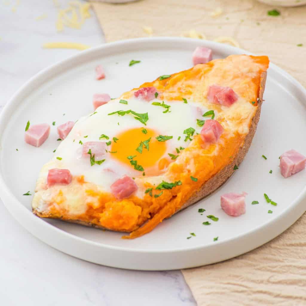 Baked sweet potato half topped with a cooked egg, melted cheese, small ham cubes, and chopped parsley, served on a white plate.