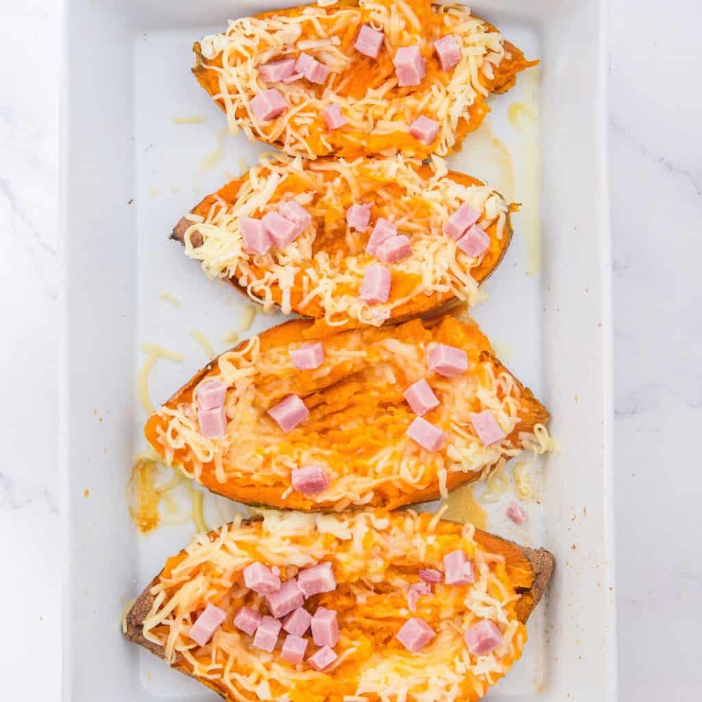 Four baked sweet potato halves topped with melted cheese and small cubes of ham are arranged in a white baking dish.