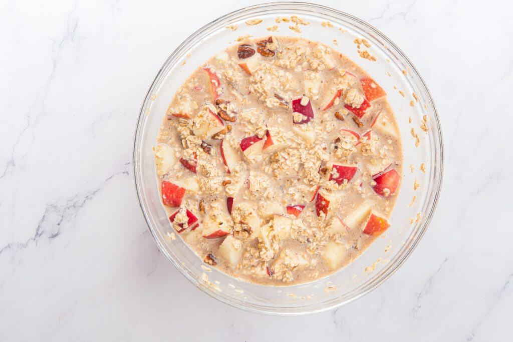 A glass bowl filled with a mixture of oats, chopped red apples, raisins, and milk sits on a white marble surface.
