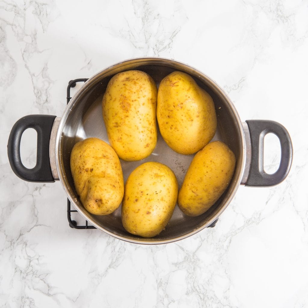 Five whole potatoes are placed inside a stainless steel pot with black handles, sitting on a marble countertop.