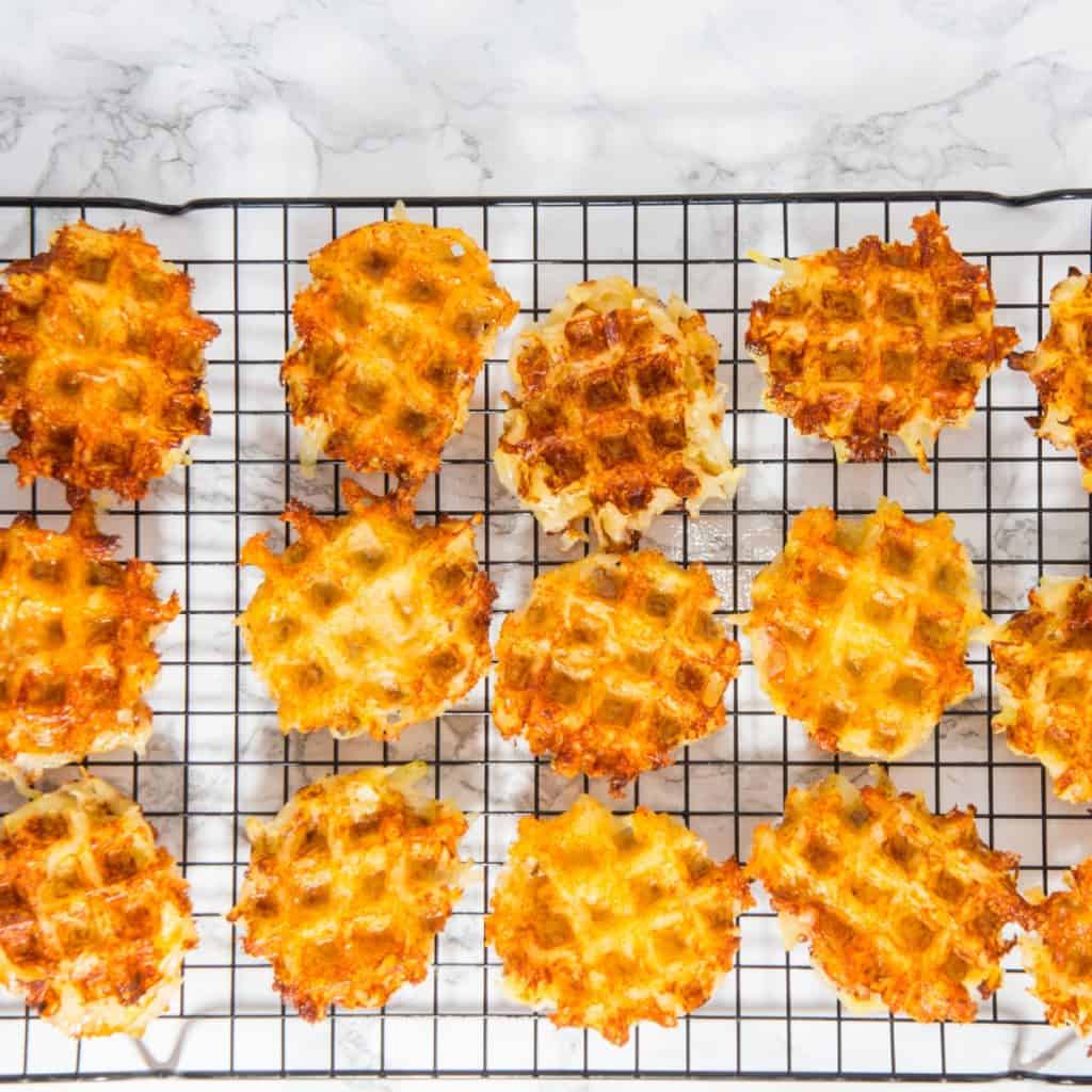 Round, golden-brown cheesy potato waffle appetizers cooling on a black wire rack, arranged in rows on a white marble surface.