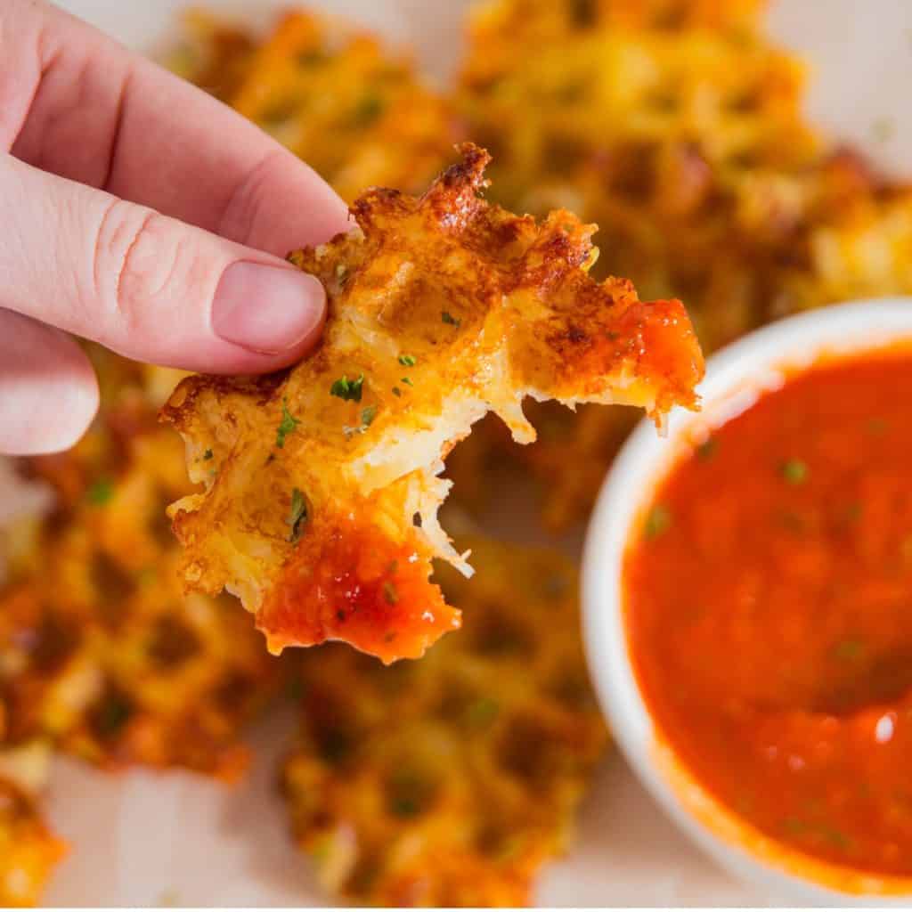 A hand holds a piece of crispy waffle with melted cheese, partially dipped in red marinara sauce. More waffles and a bowl of sauce are visible in the background.