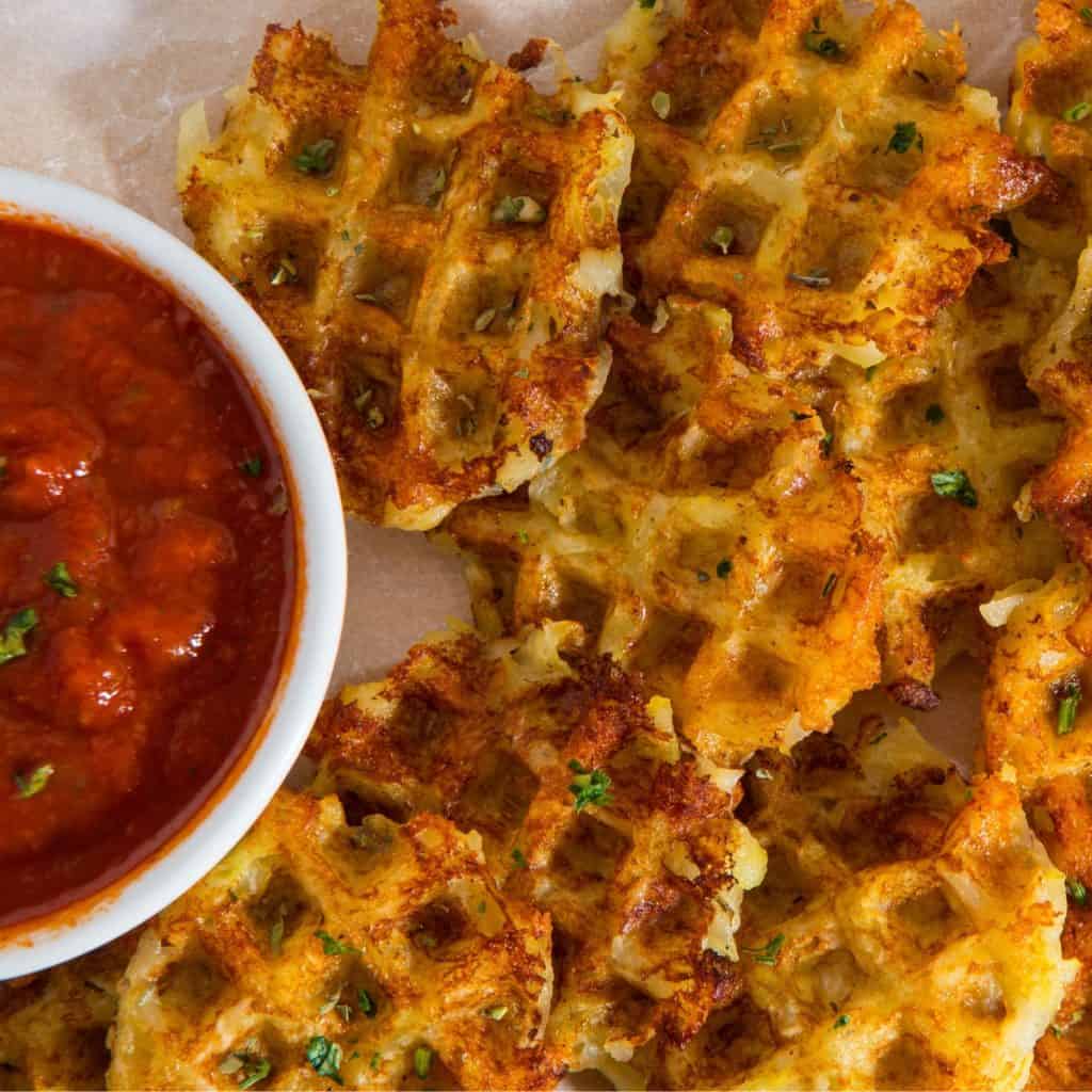 Golden-brown potato waffles garnished with herbs are arranged on parchment paper next to a bowl of chunky marinara sauce.