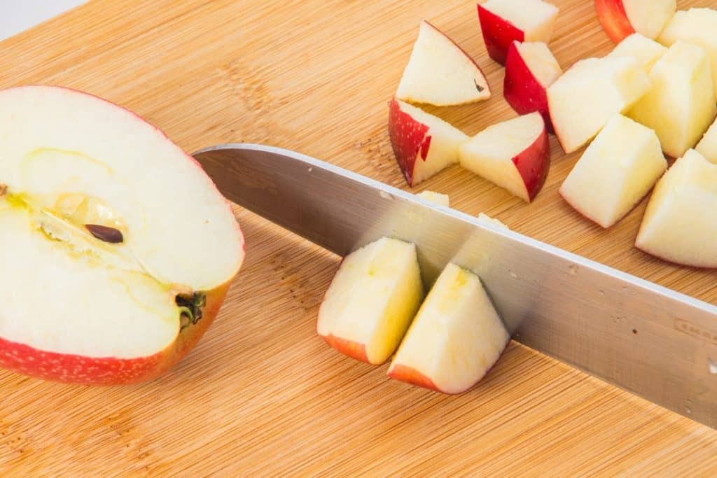 A knife slices a red apple on a wooden cutting board. Several pieces of chopped apple are visible beside the knife.