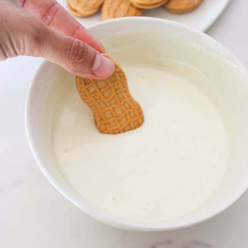 A hand dips a peanut-shaped cookie into a bowl of smooth, white cream.