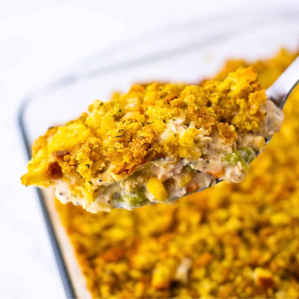 A close-up of a forkful of chicken pot pie casserole with a golden, crumbly topping and creamy filling, held above a baking dish.