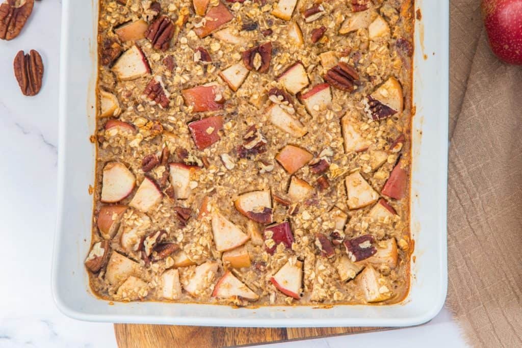 A baked oatmeal dish in a white rectangular pan, featuring visible chunks of red-skinned apples, oats, and whole pecans. A few pecans and a red apple are on the side. The scene is set on a wooden board with a beige napkin.