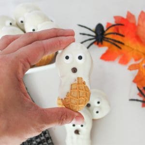 A hand holds a peanut-shaped cookie decorated as a ghost with white icing and candy eyes. Other similar cookies, an artificial spider, and an orange leaf are visible in the background.