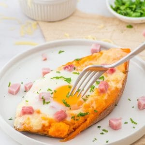 A baked sweet potato half topped with a cooked egg, melted cheese, diced ham, and chopped herbs sits on a white plate, with a fork cutting into it.