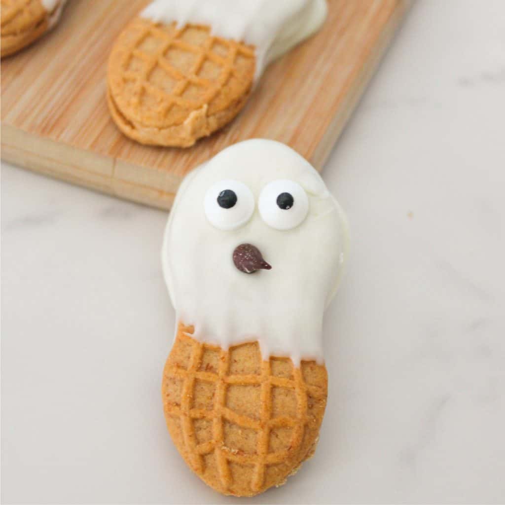 A peanut-shaped cookie partially dipped in white chocolate with candy eyes and a chocolate mouth, designed to look like a ghost, sits on a white surface near a wooden board with more cookies.