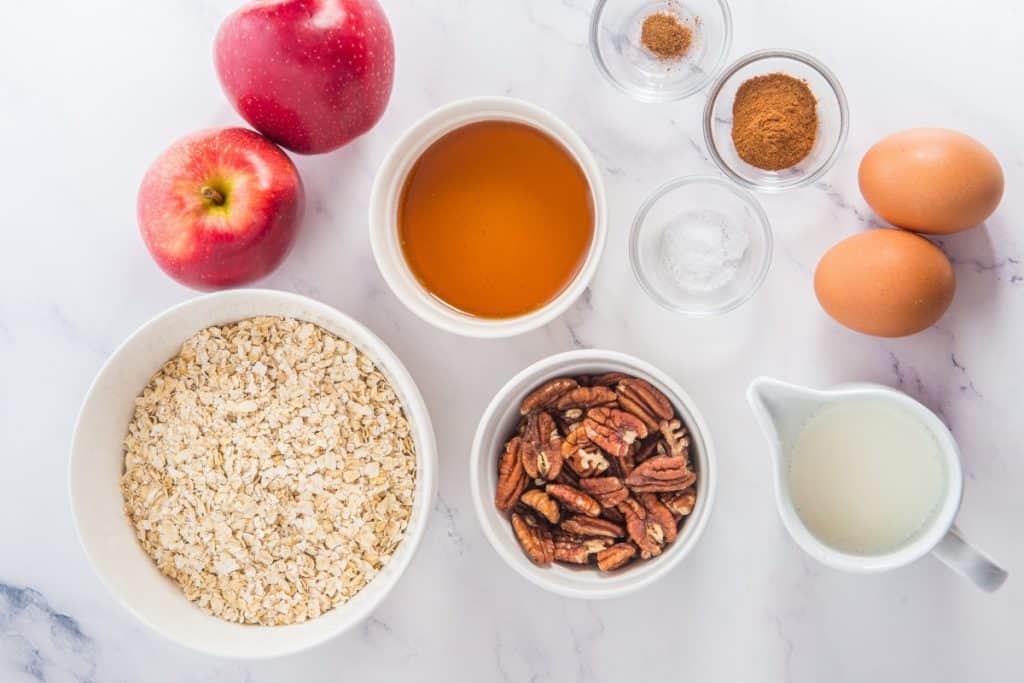 Top-down view of recipe ingredients including two apples, rolled oats, pecans, eggs, milk, honey, cinnamon, nutmeg, and salt arranged in bowls and cups on a white surface.