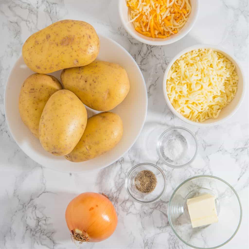 A bowl of raw potatoes, an onion, grated cheddar cheese, grated mozzarella cheese, butter, and small bowls of salt and pepper are arranged on a white marble surface.