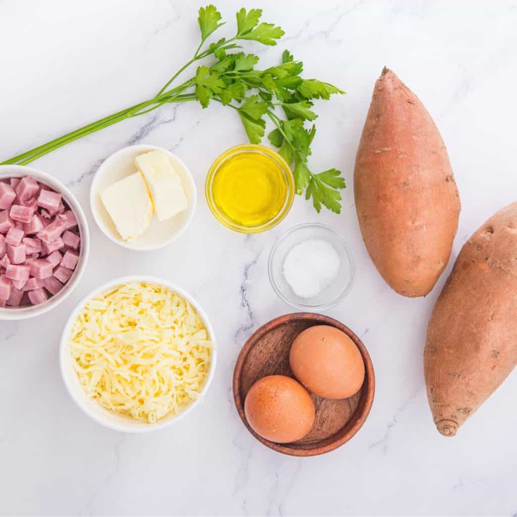 Two whole sweet potatoes, two eggs, chopped ham, shredded cheese, butter, olive oil, salt, and parsley arranged on a white marble surface.