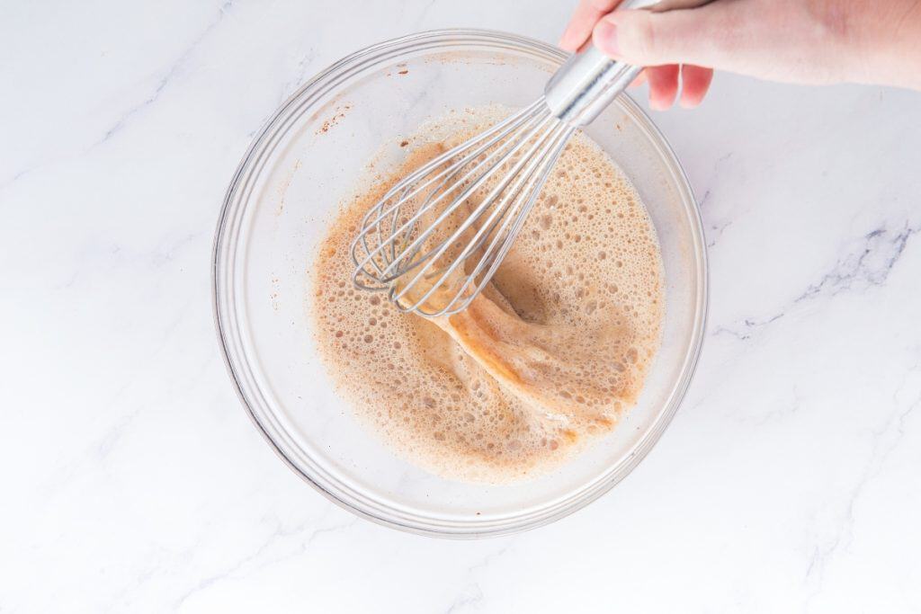 A hand holds a metal whisk, mixing a light brown, frothy liquid in a clear glass bowl on a white marble surface.