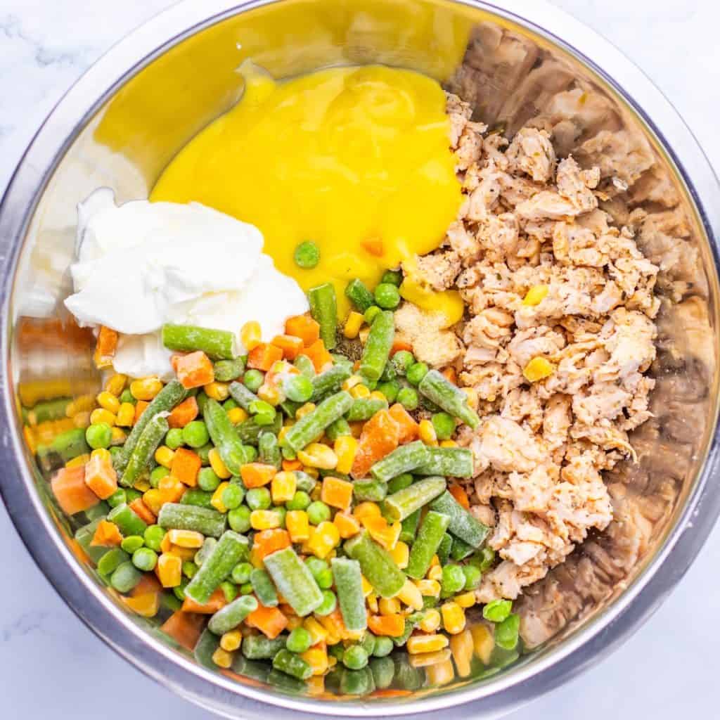 Mixing the ingredients for chicken stuffing pot pie casserole, a metal bowl holds flaked tuna, frozen mixed vegetables, cream of chicken soup, and a scoop of sour cream before blending together.