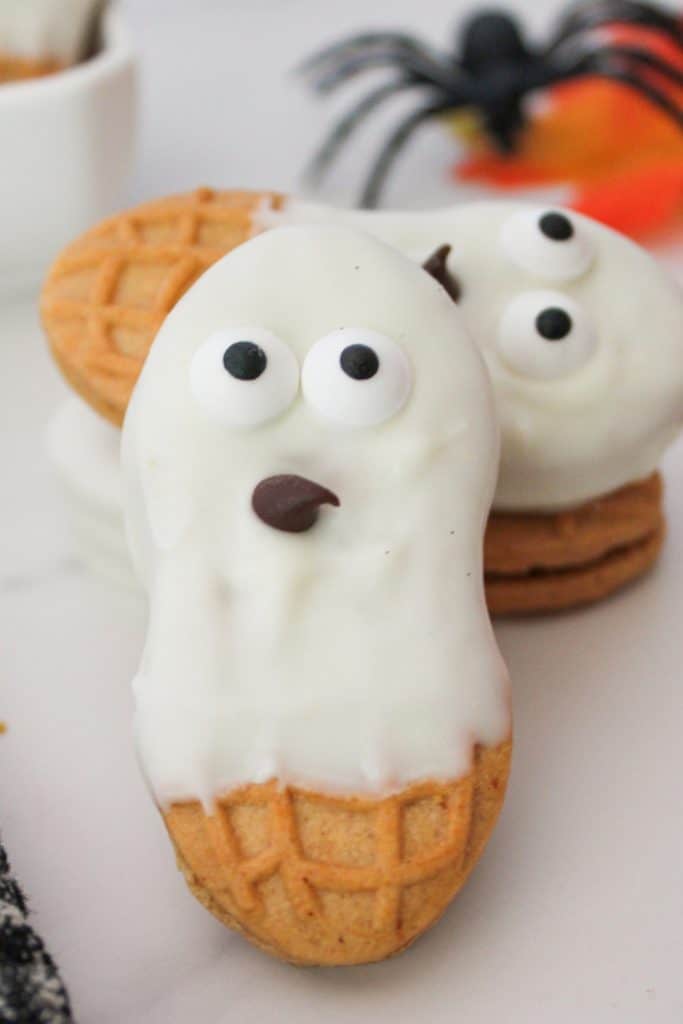 A peanut-shaped cookie partially dipped in white chocolate, decorated with candy eyes and a chocolate chip mouth to resemble a ghost, with other similar cookies and a plastic spider in the background.