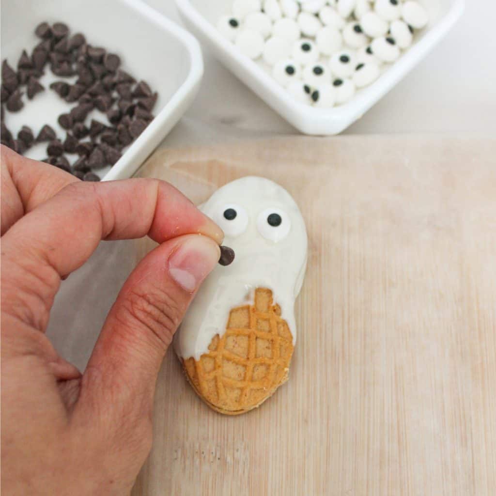 A hand places a chocolate chip on a ghost-shaped cookie with white icing and candy eyes. Bowls of chocolate chips and candy eyes are nearby.