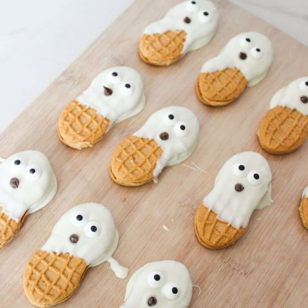 Nutter Butter cookies partially dipped in white chocolate to resemble ghosts, with candy eyes and chocolate chip mouths, arranged on a wooden surface.