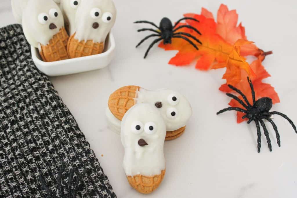 Cookies dipped in white frosting with candy eyes and chocolate mouths arranged to look like ghosts, placed on a white surface with two black fake spiders, orange autumn leaves, and a black textured cloth.