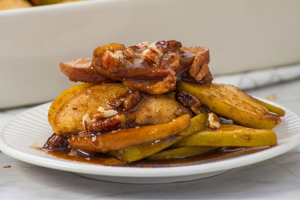Sliced baked apples with cinnamon, pecans, and a brown sauce are stacked on a white plate.