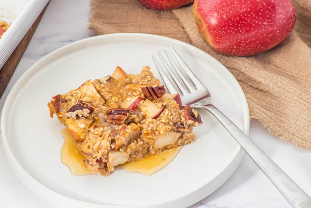 A square serving of baked oatmeal with apple chunks and pecans sits on a white plate with a fork, drizzled with syrup. Two red apples and a brown cloth are visible in the background.