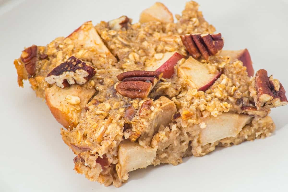A square slice of baked oatmeal with visible apple pieces and pecan halves sits on a plain white plate.