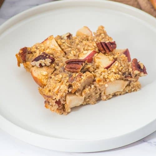 A square slice of oatmeal bake with visible apple chunks and pecan pieces sits on a round white plate, with part of a red apple and a baking dish blurred in the background.