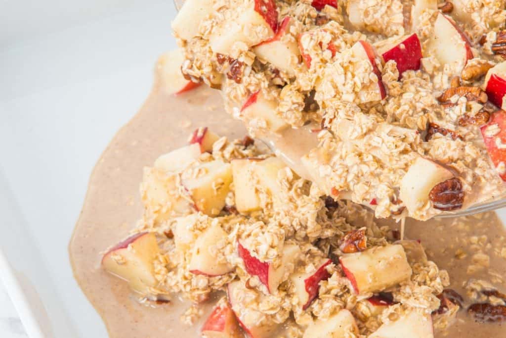 Chopped apples and oats mixed with a creamy sauce are being poured from a bowl into a dish, creating a thick, textured mixture.