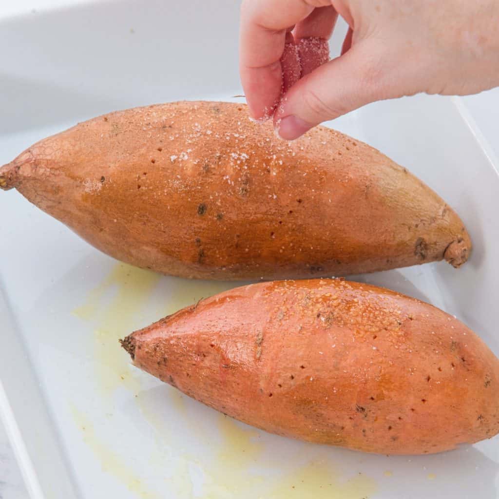 A hand sprinkles salt over two whole sweet potatoes placed in a white baking dish, with some oil visible on the surface.
