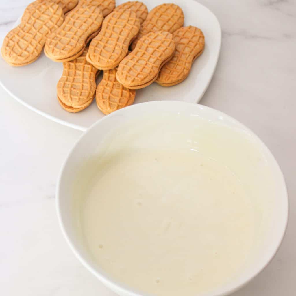 A white bowl filled with melted white chocolate sits next to a white plate with several peanut-shaped sandwich cookies on a marble surface.