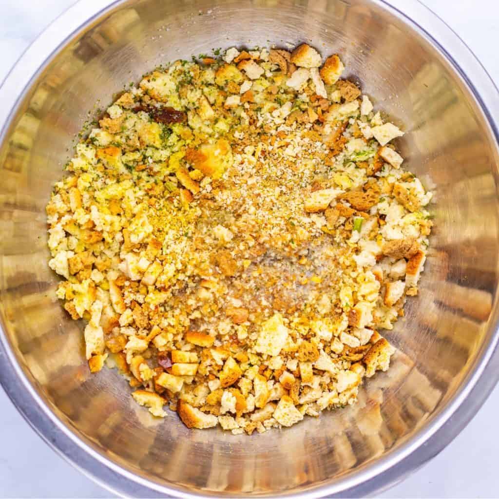 A stainless steel mixing bowl containing crushed bread cubes, dried herbs, and seasoning, likely for making stuffing or dressing.