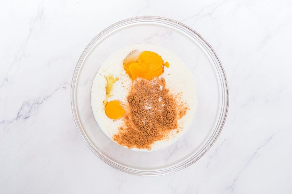 Clear glass bowl containing flour, two cracked eggs, a heap of ground cinnamon, and a white liquid, all unmixed, on a white marble surface.