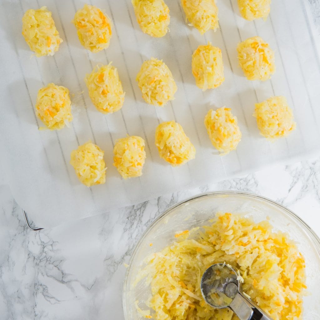 Rows of uncooked potato and cheese mixture balls are arranged on a parchment-lined rack. A glass bowl with leftover mixture and a metal scoop is visible at the bottom right on a marble surface.